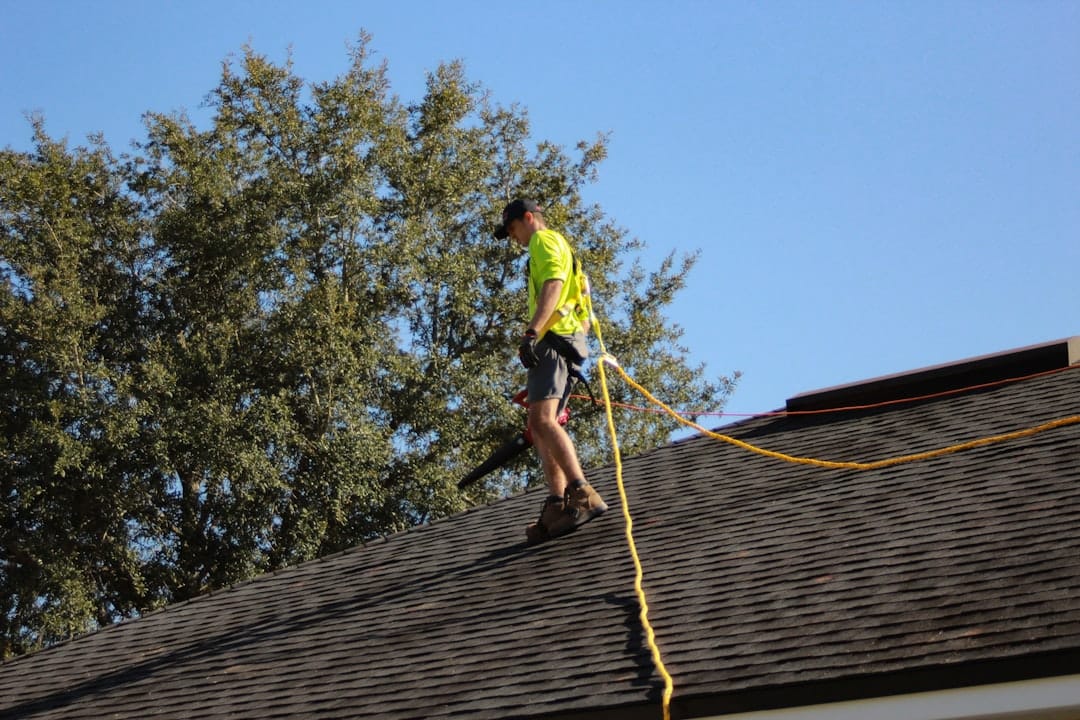 Professional roofer wearing a safety harness while using a leaf blower to clear debris during roof maintenance in Leon Valley TX
